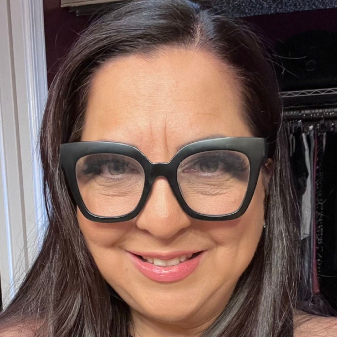 Woman wearing large black glasses and smiling, with long dark hair and a closet in the background.