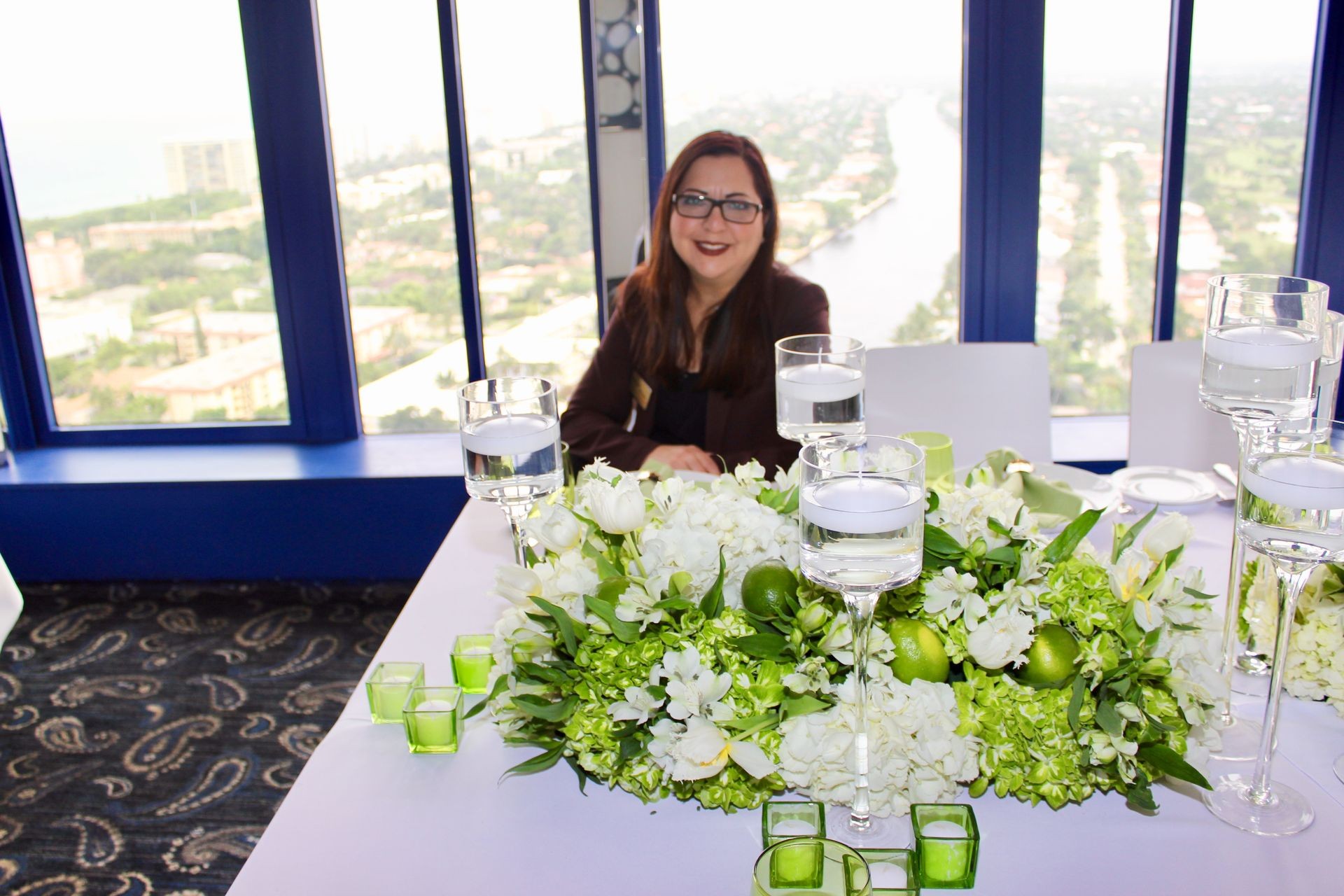 Woman sitting at a white table with floral centerpiece and candle holders, overlooking a cityscape view.