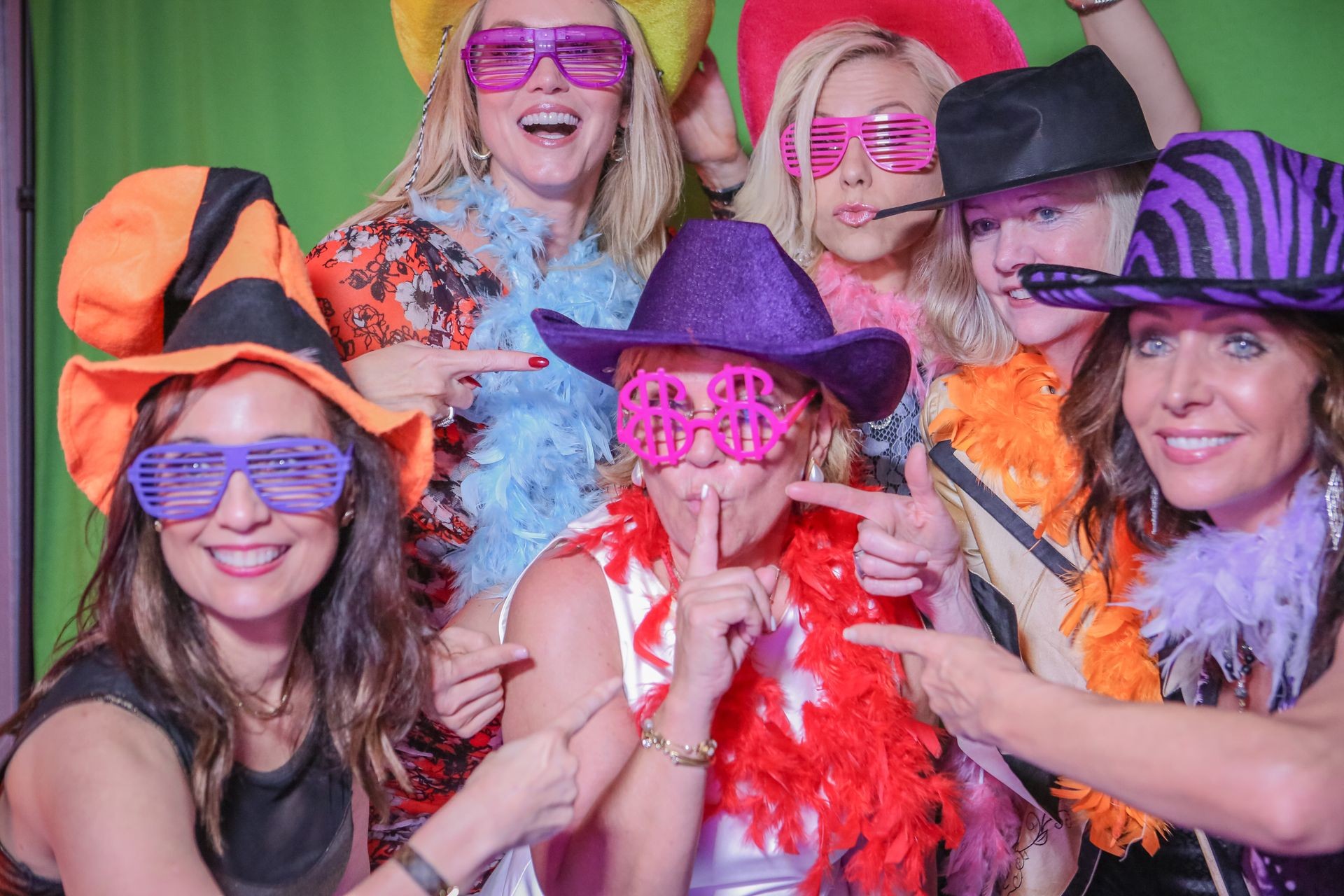 Group of women wearing colorful hats, feather boas, and novelty glasses posing playfully.